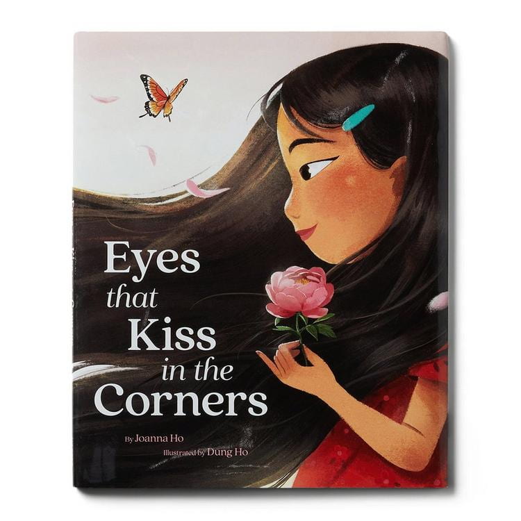 An image of the book cover of Eyes that Kiss in the Corners by Joanna Ho and illustrated by Dung Ho. There is an illustration of an Asian child with long black hair in profile. They are holding a pink rose and looking at a butterfly. They have a blue clip in their hair and they're wearing a red shirt.